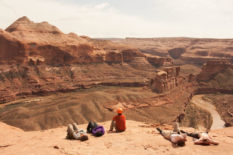The image shows a scenic view of a canyon landscape with a group of people relaxing on the edge. The canyon features layered red rock formations and a river winding through the bottom. The people are casually positioned, some lying down and others sitting, enjoying the view. The overall impression is one of relaxation and appreciation of the natural beauty.